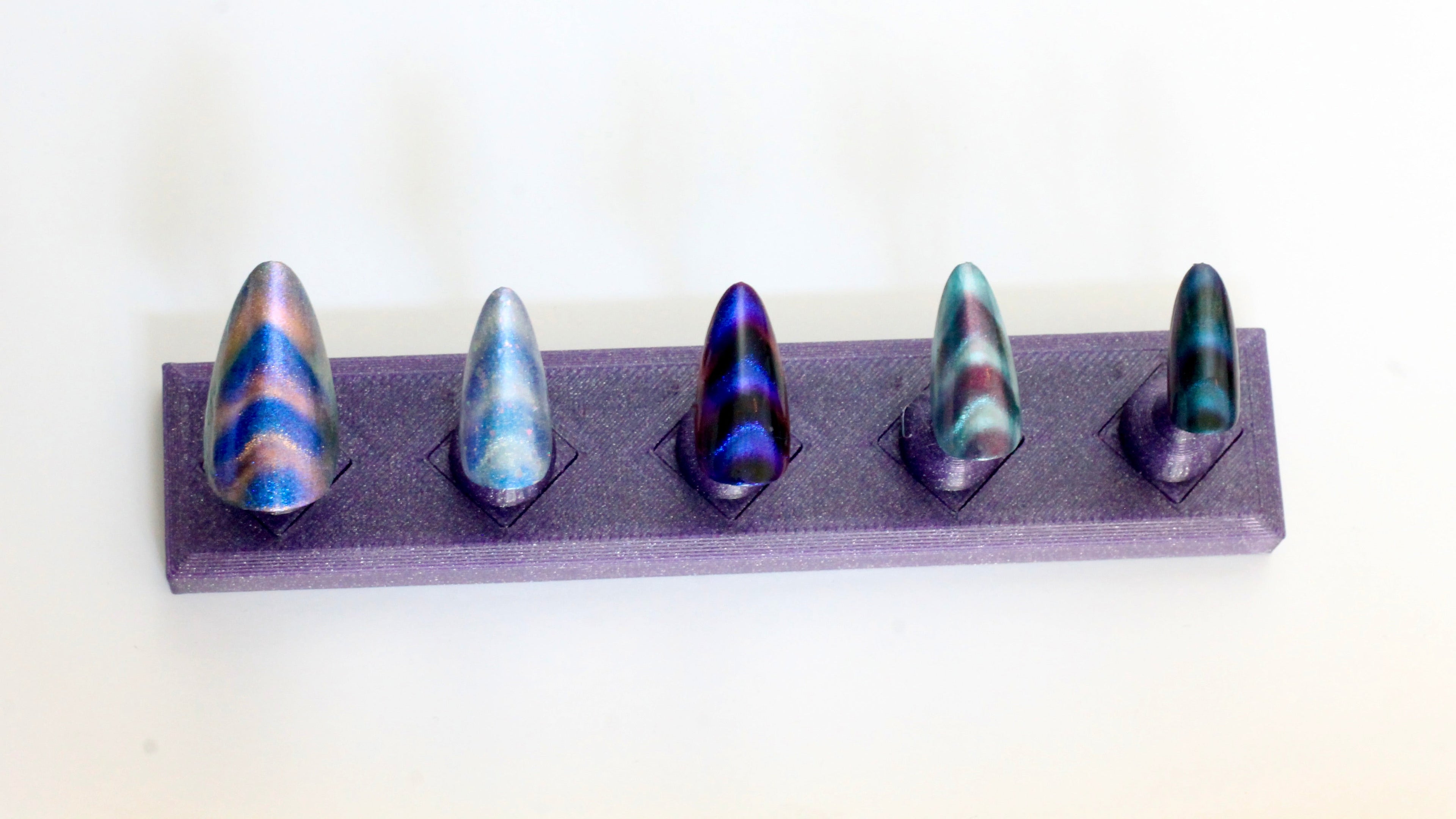 Five press-on nails painted with magnetic metallic polish of various colors on a purple stand, against a white background